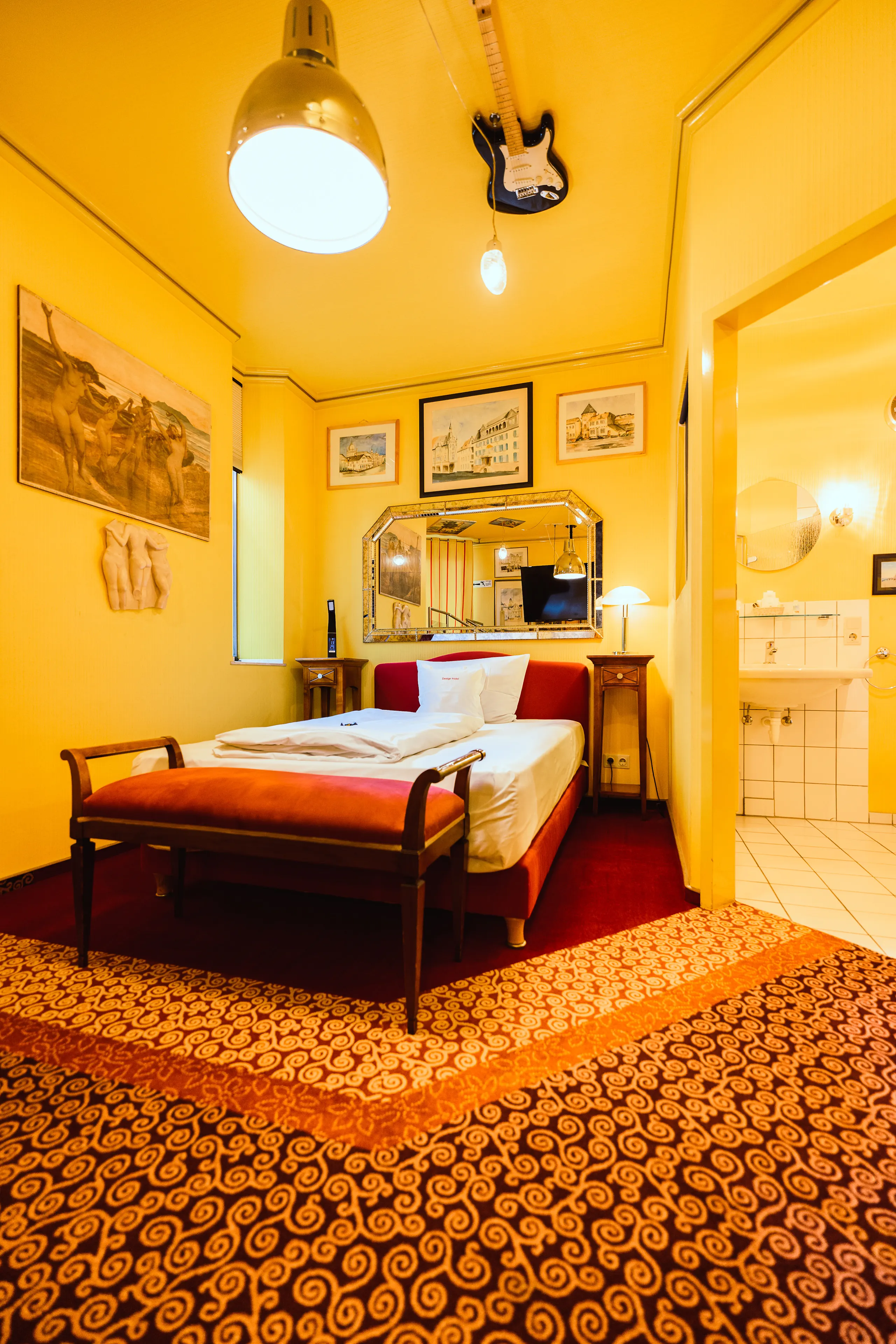 Design themed room in yellow with a single bed, a red bench and a guitar mounted on the ceiling.